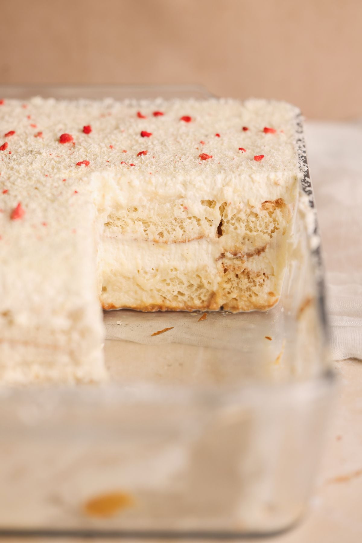 A glass baking dish filled with a creamy layered rus malai tiramisu, topped with white cream and grated white chocolate, with a large piece removed to reveal the fluffy, airy interior layers.