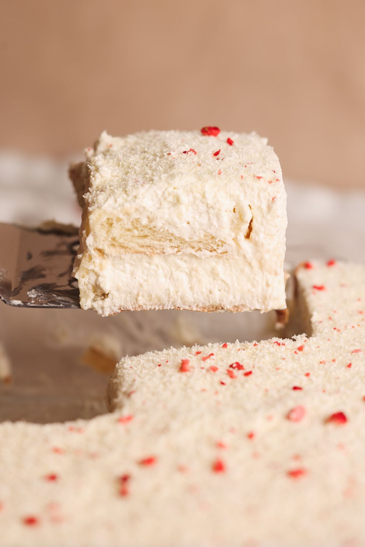 A close-up of a creamy, layered rus malai tiramisu slice with white topping, lifted by a spatula above the rest of the dessert.