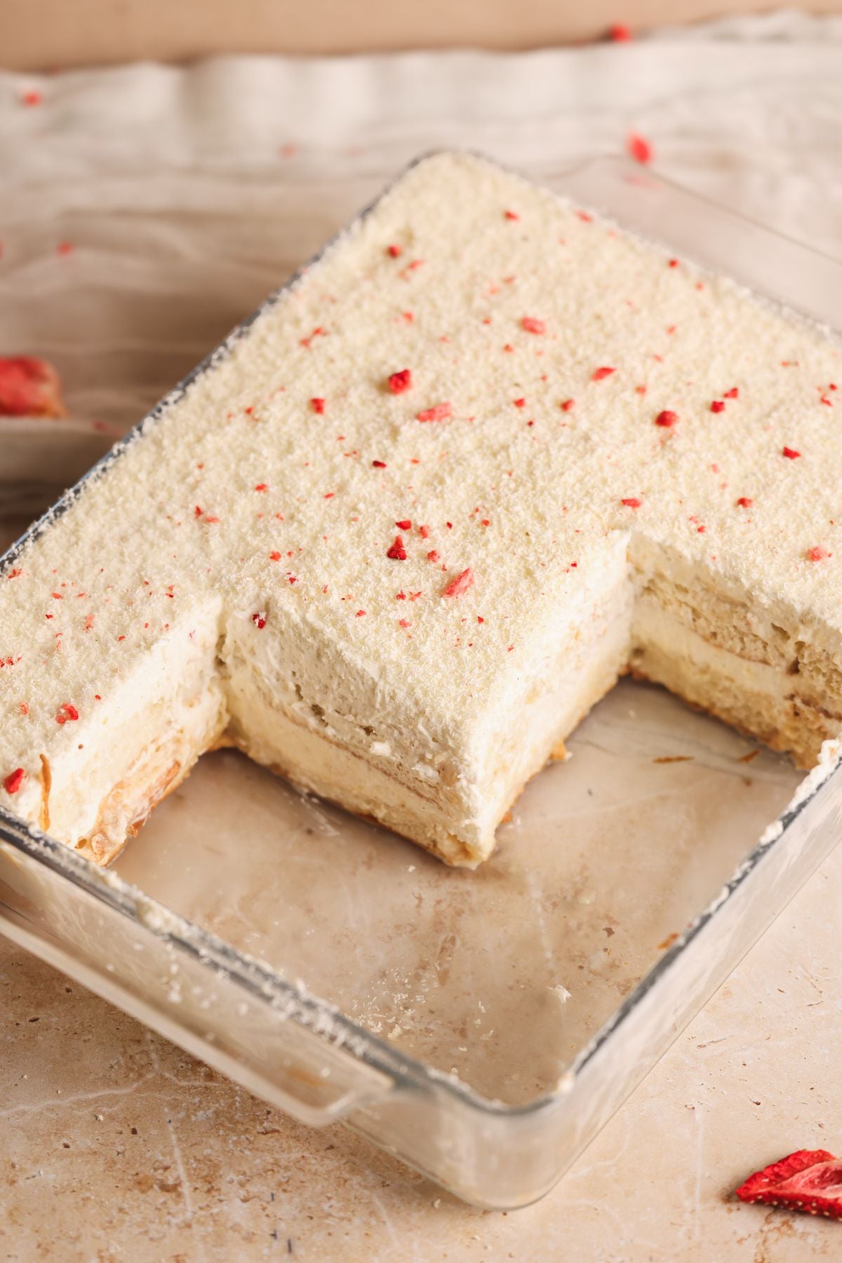 A rectangular glass dish holds a layered rus malai tiramisu with a creamy, pale filling and a white chocolate topping sprinkled with small red crushed strawberries. A large square piece has been removed, revealing the dessert’s layers.
