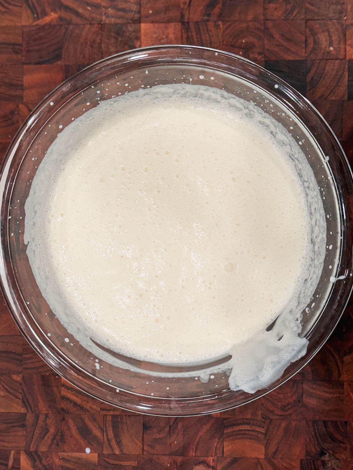 A clear glass bowl filled with a creamy, frothy semi whipped cream sits on a dark wooden surface. Some batter is slightly splashed on the rim of the bowl.