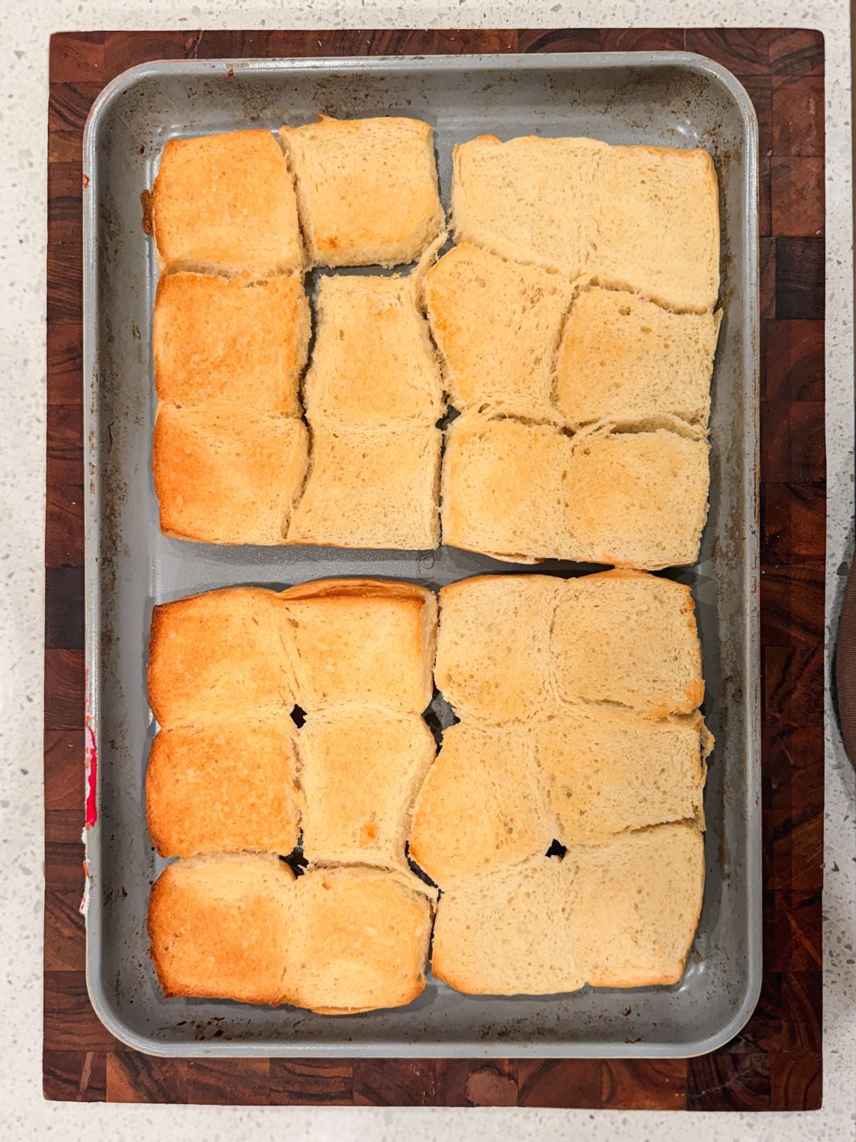 A metal baking tray holds twelve square bread rolls, arranged in four rows of three. The bread is lightly toasted, with some pieces more golden brown than others. The tray sits on a dark wooden surface.