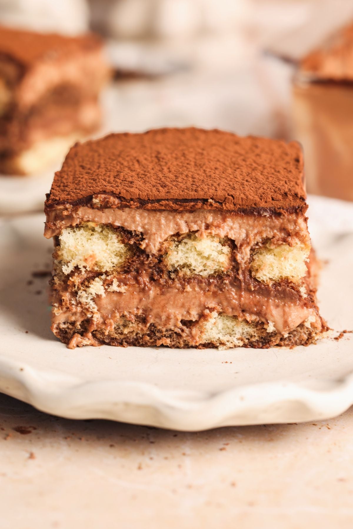 A close-up of a slice of triple chocolate tiramisu dessert on a white plate, showing layers of chocolate cream, soaked ladyfingers, and cocoa powder dusted on top.
