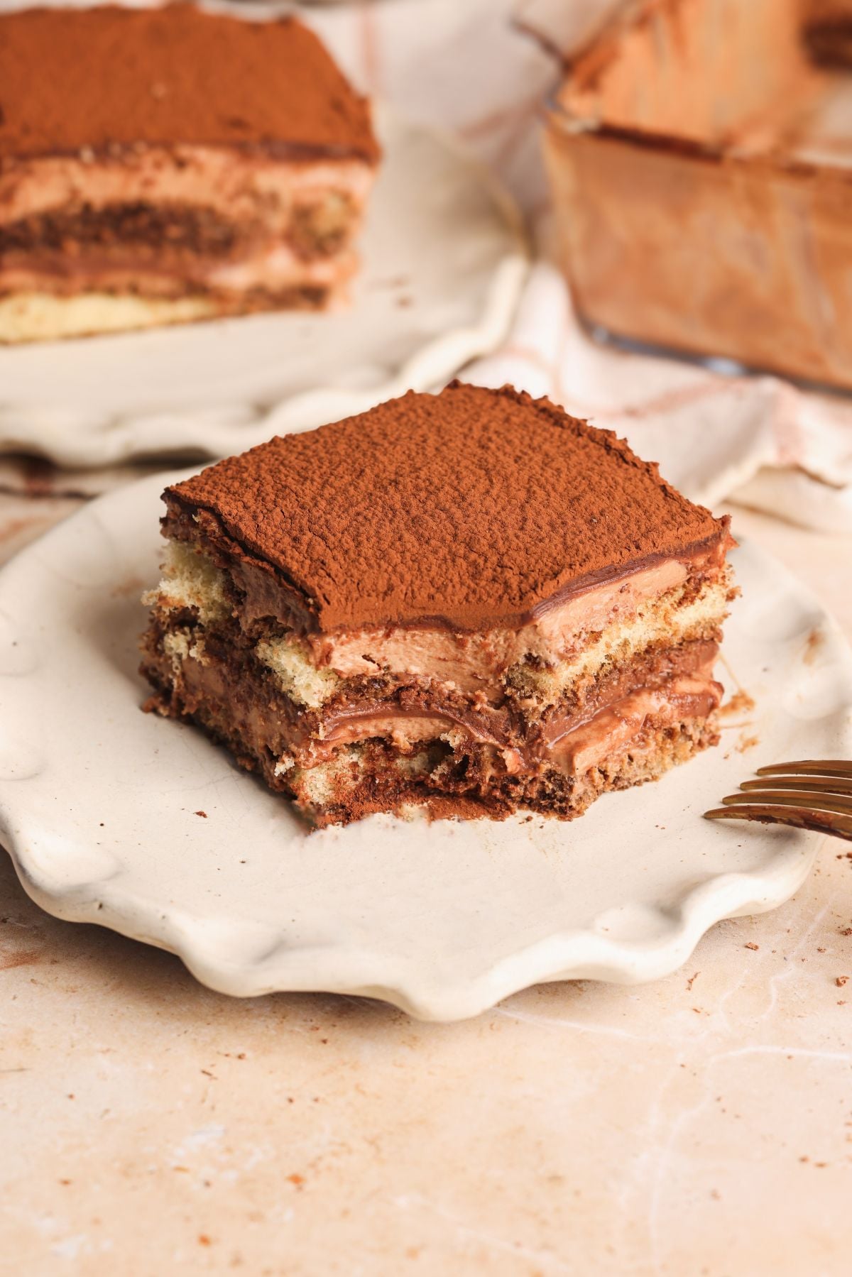 A slice of triple chocolate tiramisu with visible creamy and layers, dusted with cocoa powder, sits on a white wavy-edged plate with a fork nearby. Another slice is in the background.