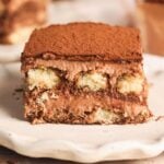A close-up of a slice of chocolate tiramisu on a white plate, showing layers of coffee-soaked ladyfinger biscuits and creamy chocolate mascarpone filling, topped with a dusting of cocoa powder.