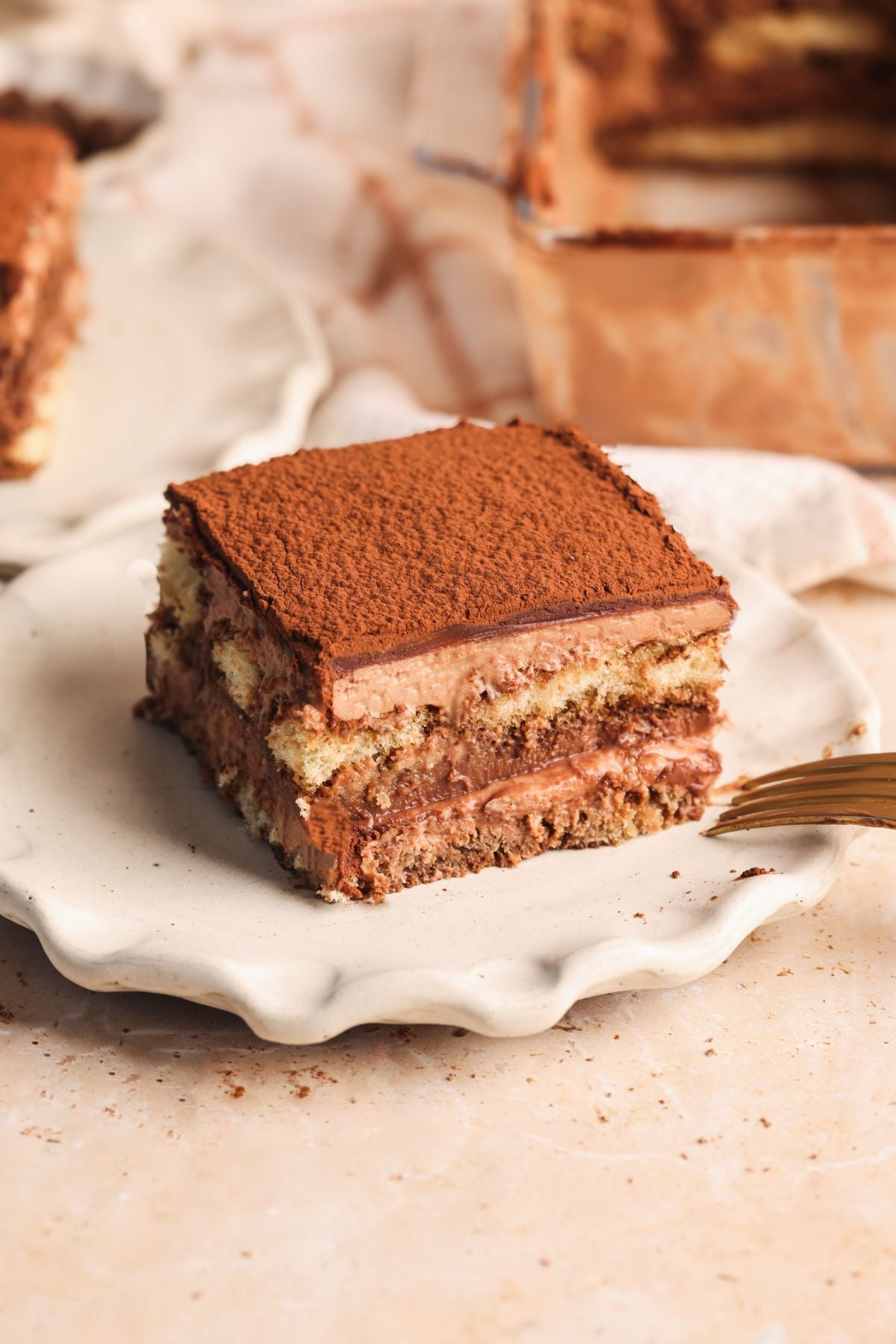 A square slice of triple chocolate tiramisu sits on a white plate, showing layers of coffee-soaked ladyfingers and creamy filling, topped with a dusting of cocoa powder. A gold fork rests nearby on the light-colored surface.