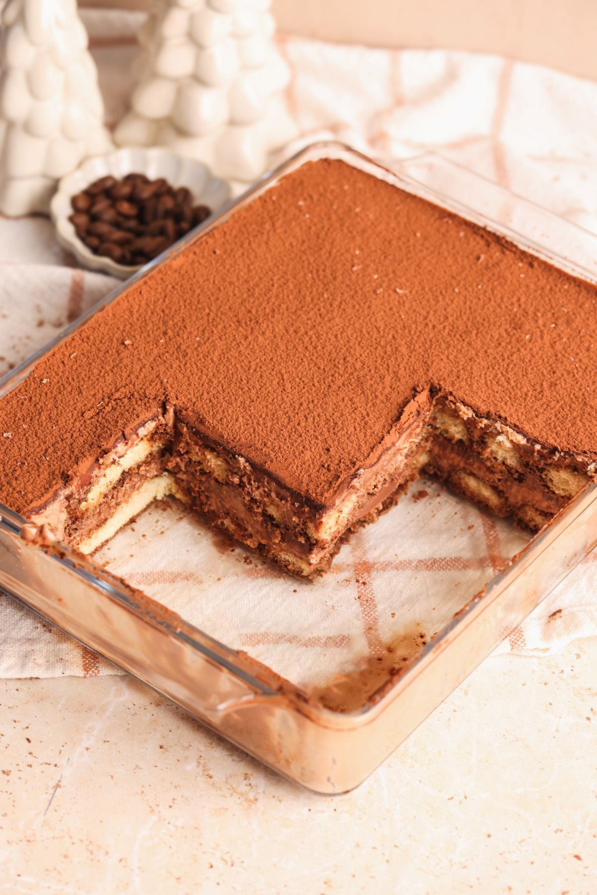 A glass dish of chocolate tiramisu with a large square piece removed, showing layers of cream and coffee-soaked ladyfingers. The dessert is topped with cocoa powder, and there are white ceramic trees and a small bowl of coffee beans in the background.