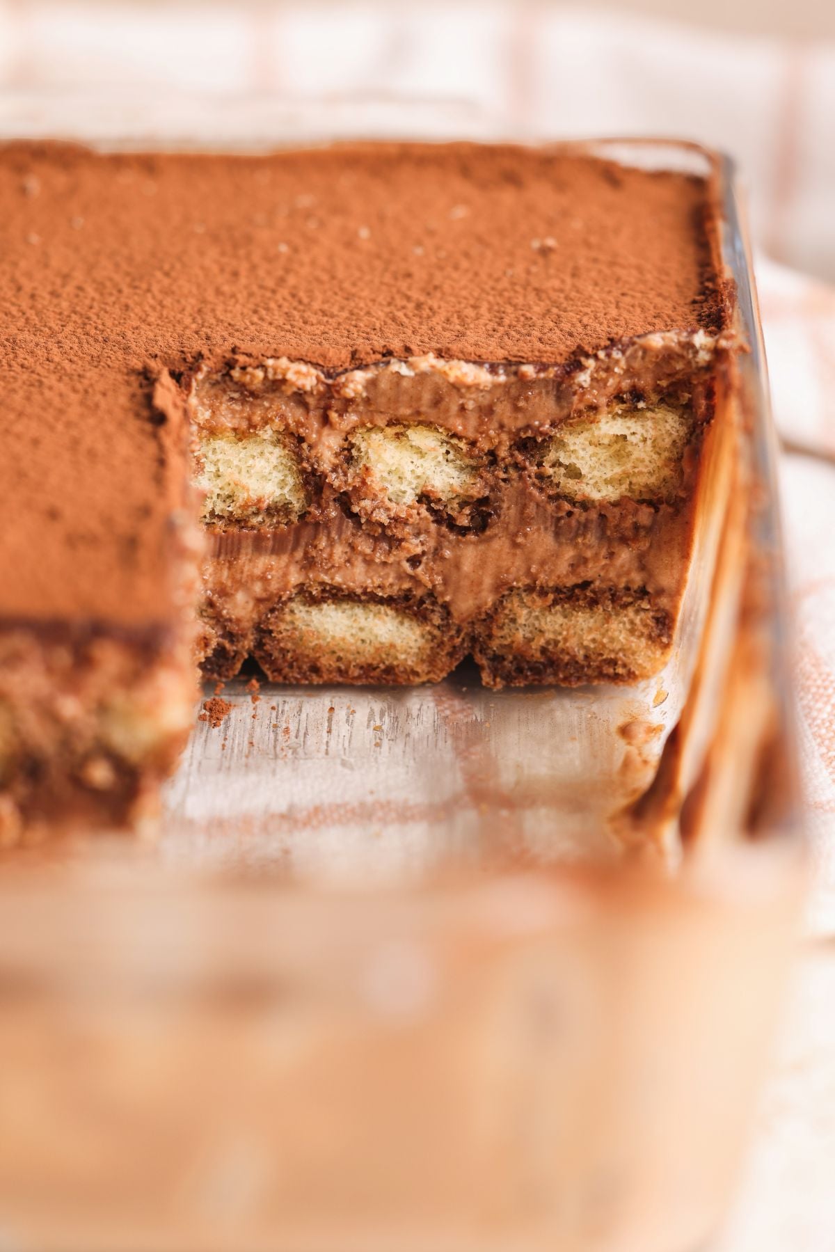 A close-up of a glass dish filled with triple chocolate tiramisu, showing layers of coffee-soaked ladyfingers and creamy chocolate mascarpone filling, topped with a dusting of cocoa powder; a portion has been scooped out.