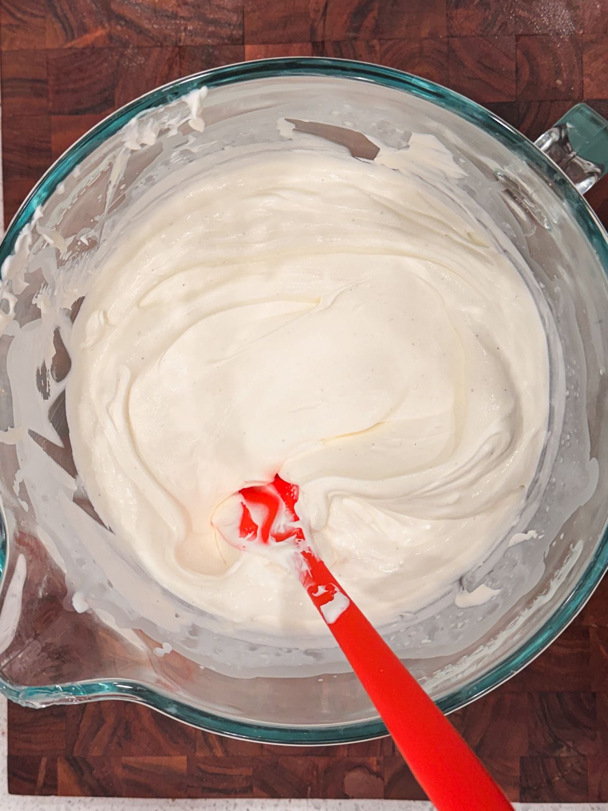 A glass bowl filled with creamy, whipped cream cheese frosting and a red spatula, sitting on a dark wooden surface.