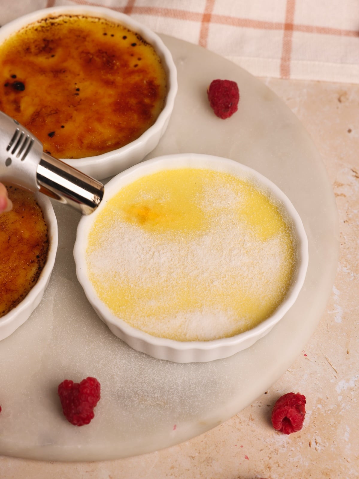 A hand uses a kitchen blow torch to caramelize sugar on top of a crème brûlée in a white ramekin. Two finished crème brûlées and some raspberries are nearby on a round marble surface.