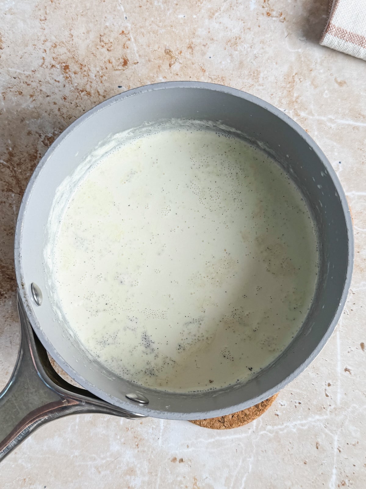A saucepan filled with a simmering creamy mixture, speckled with vanilla beans, sits on a light-colored countertop. The mixture appears to be gently heated, possibly for a dessert recipe.