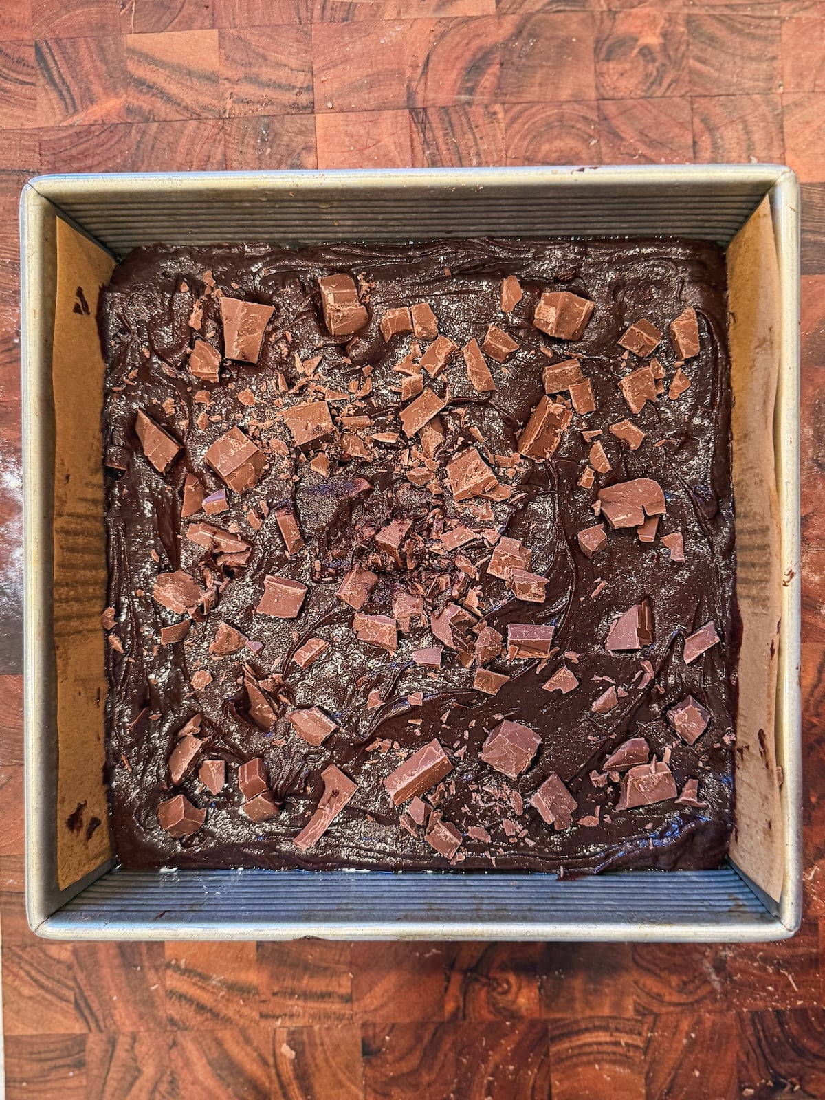 A square metal baking pan filled with chocolate brownie batter, topped with chunks of chocolate, sits on a wooden countertop.
