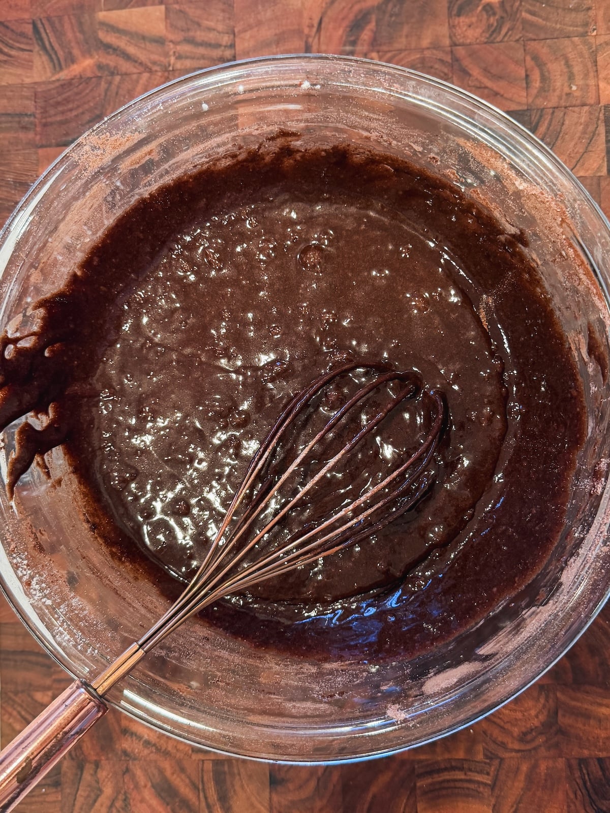 A glass bowl filled with chocolate brownie batter is being mixed with a metal whisk, resting on a wooden countertop.