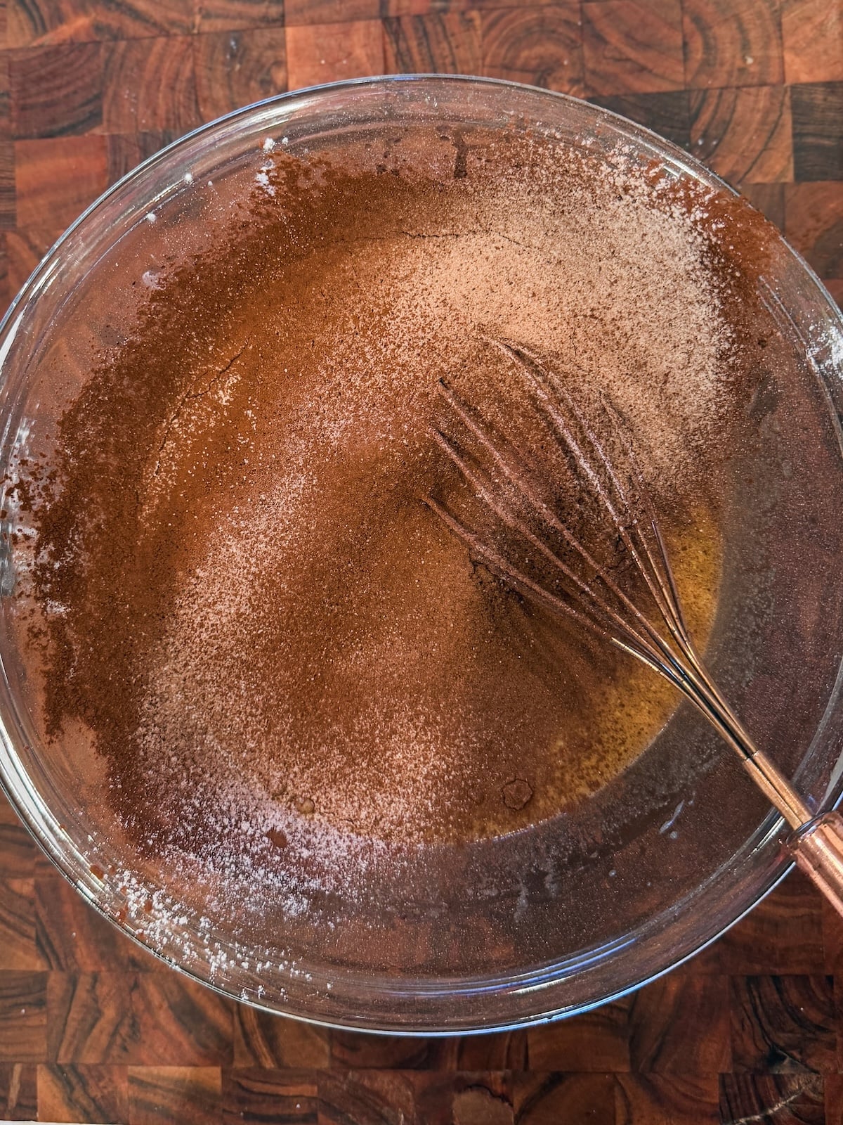 A glass bowl filled with ingredients being mixed, including cocoa powder and flour, with a whisk resting inside on a wooden countertop.