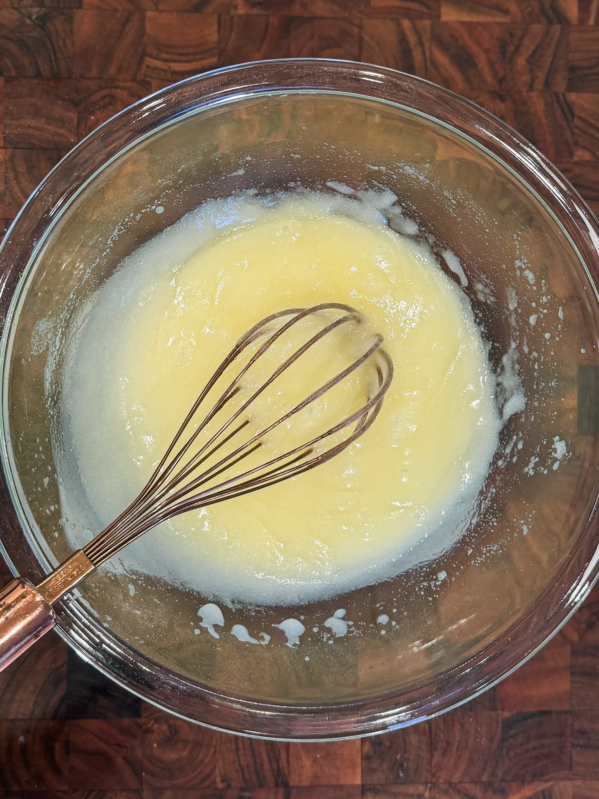 A metal whisk rests in a glass bowl containing a pale yellow mixture of sugar and butter on a wooden countertop.