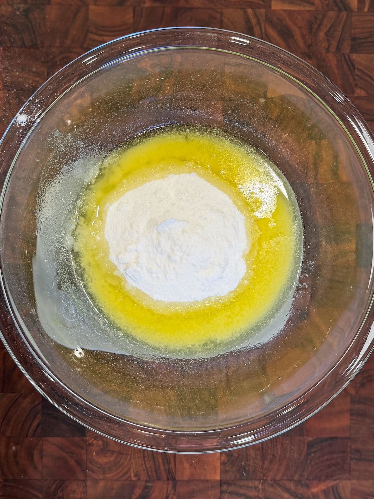 A glass mixing bowl on a wooden surface, containing melted butter and a mound of sugar in the center, ready to be mixed together.
