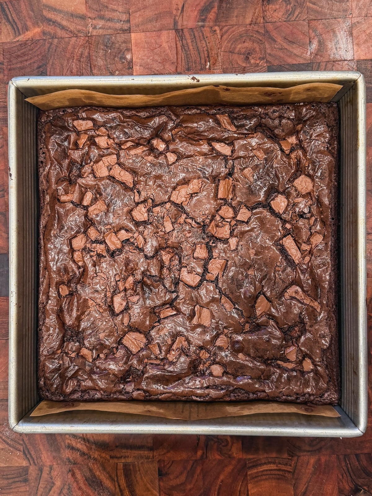 A square baking pan filled with freshly baked chocolate brownies, featuring a shiny, crinkled top and scattered chunks of chocolate. The pan rests on a wooden surface.