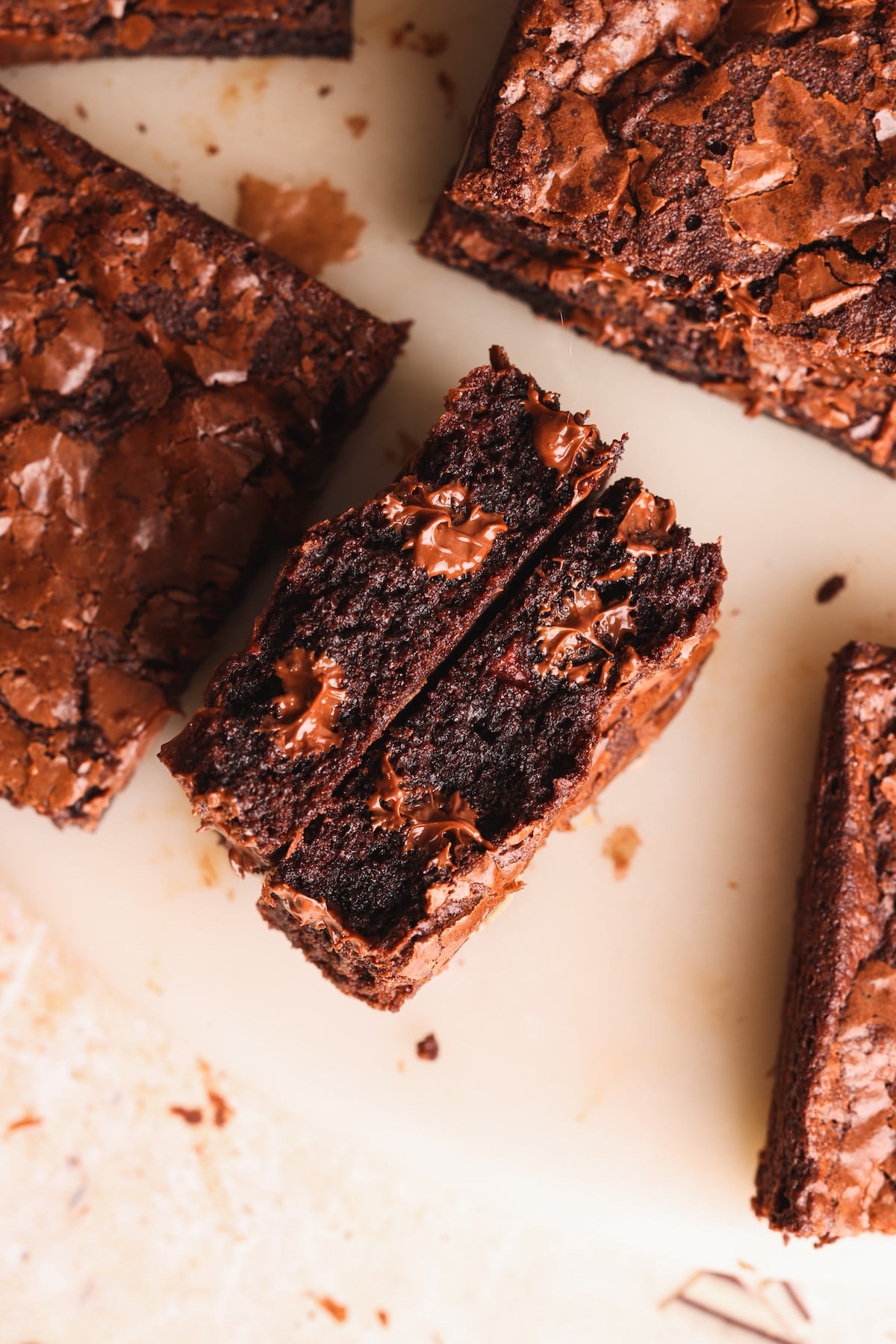 Close-up of rich chocolate brownies on a light surface, with one brownie split in half to reveal melted chocolate chips inside and a glossy, crackly top.