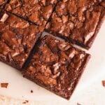Close-up of several chewy, fudgy chocolate brownies with a shiny, crackled top, arranged on a round white plate. The brownies are cut into neat squares and have a rich, chocolatey appearance.