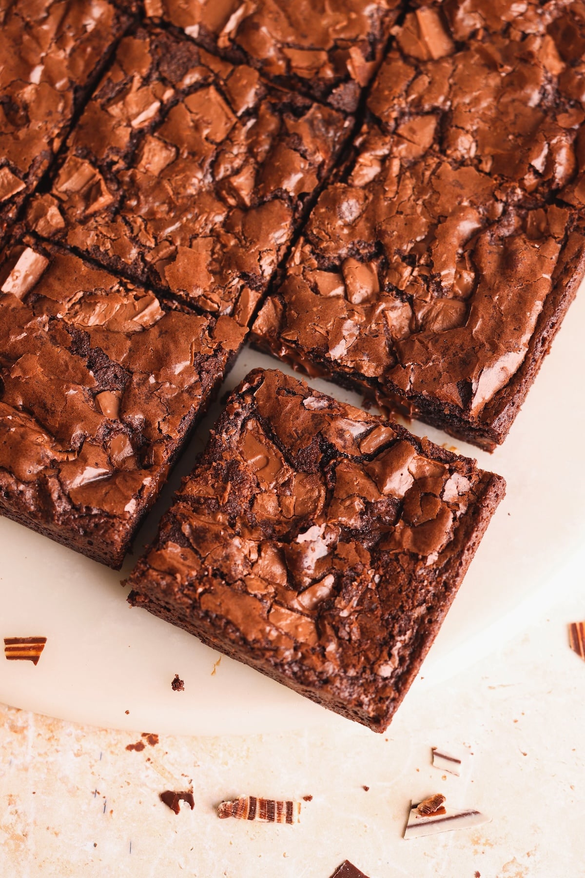 A close-up of chocolate brownies sliced into squares on a white surface, with a rich, cracked top and chocolate chunks visible. One brownie piece is slightly pulled out from the rest.