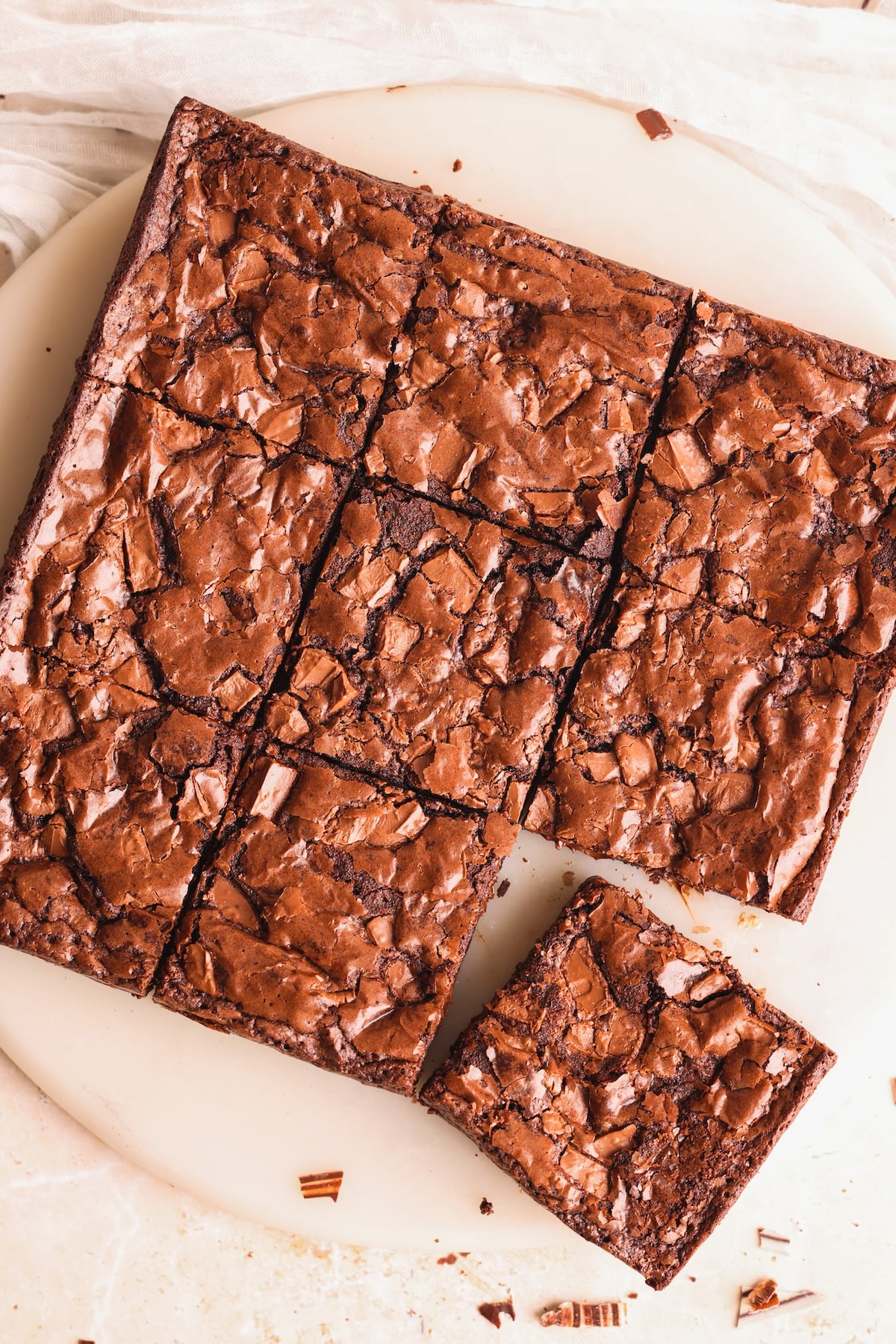 A batch of chocolate brownies with a shiny, crackled top, cut into nine squares on a round white plate. One brownie square is slightly separated from the rest.