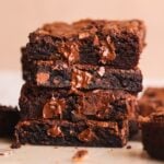 A close-up of a stack of three rich, fudgy chocolate brownies with melted chocolate chunks oozing out, set against a light background. Another brownie is blurred in the background.