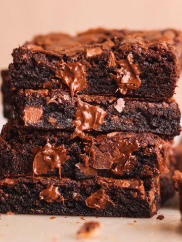 A close-up of a stack of three rich, fudgy chocolate brownies with melted chocolate chunks oozing out, set against a light background. Another brownie is blurred in the background.