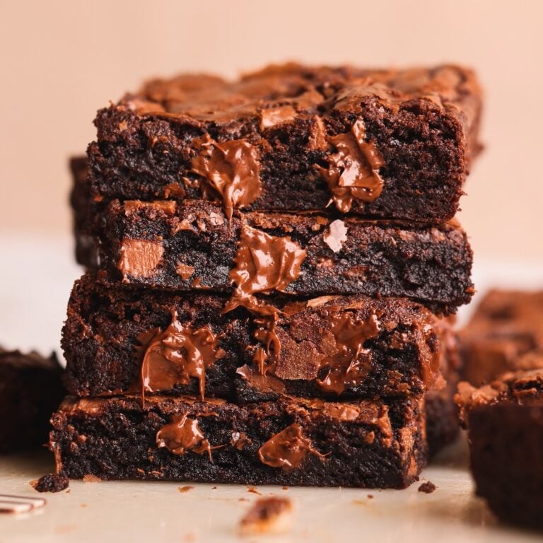 A close-up of a stack of three rich, fudgy chocolate brownies with melted chocolate chunks oozing out, set against a light background. Another brownie is blurred in the background.