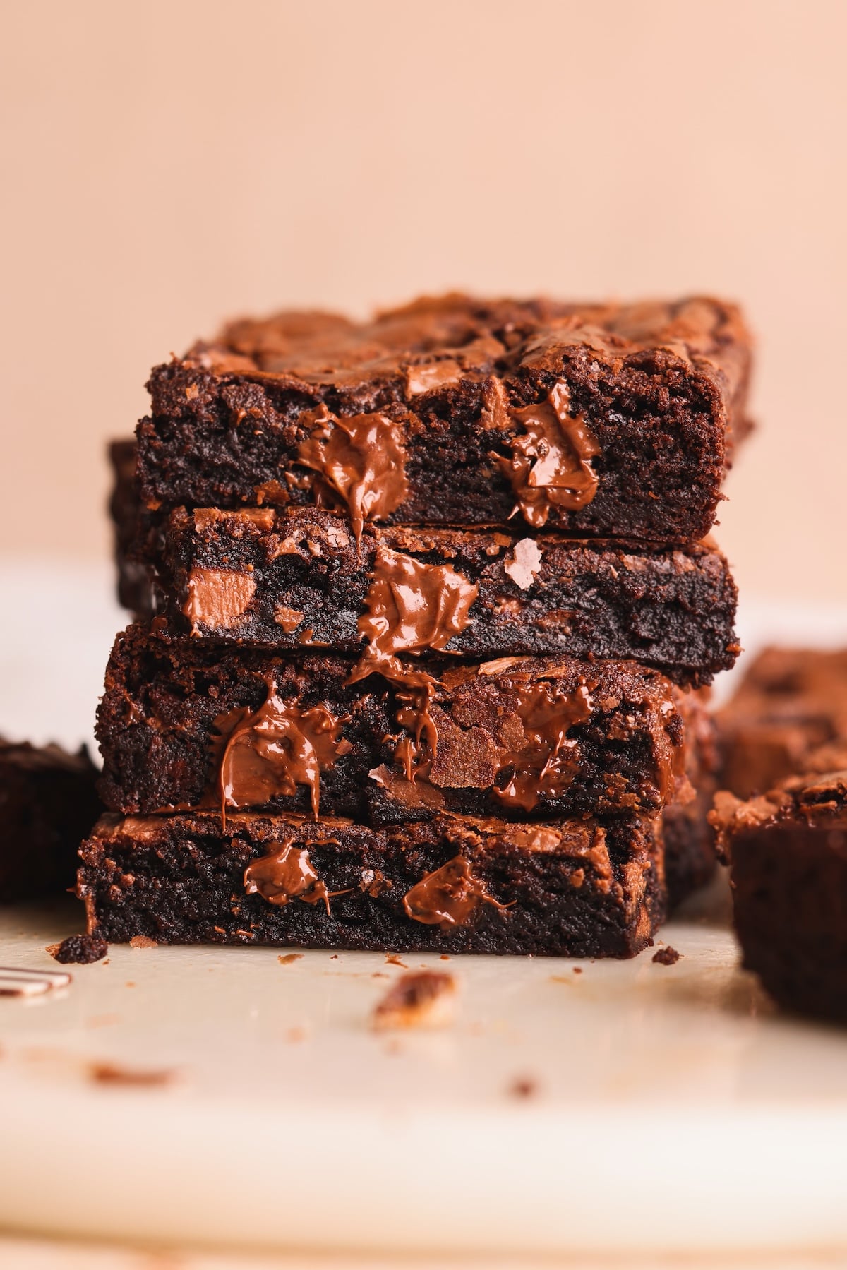 A close-up of a stack of thick, fudgy brownies with melted chocolate chunks, resting on a light-colored surface. The brownies appear rich, moist, and freshly baked.