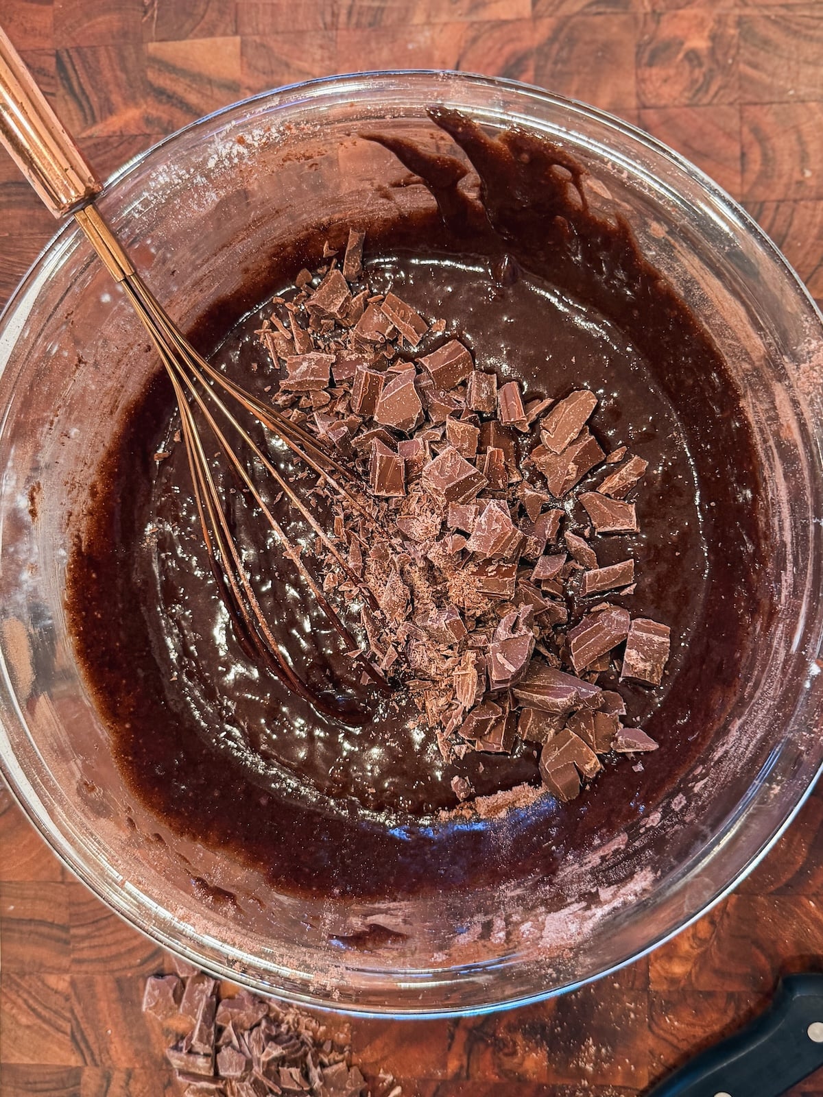A glass bowl filled with chocolate brownie batter and chunks of chopped chocolate, being mixed with a metal whisk, sits on a wooden surface.