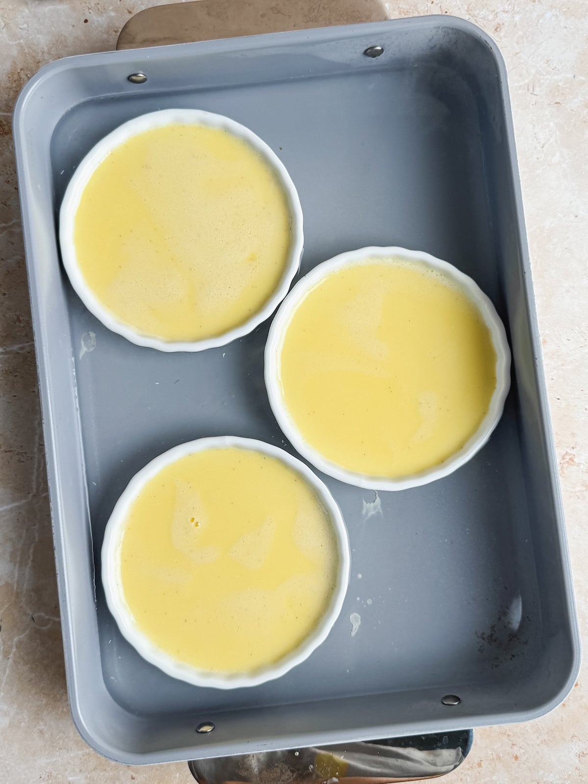 Three white ramekins filled with a pale yellow custard mixture are placed in a large gray baking pan with boiling water on a light-colored countertop, ready to be baked.