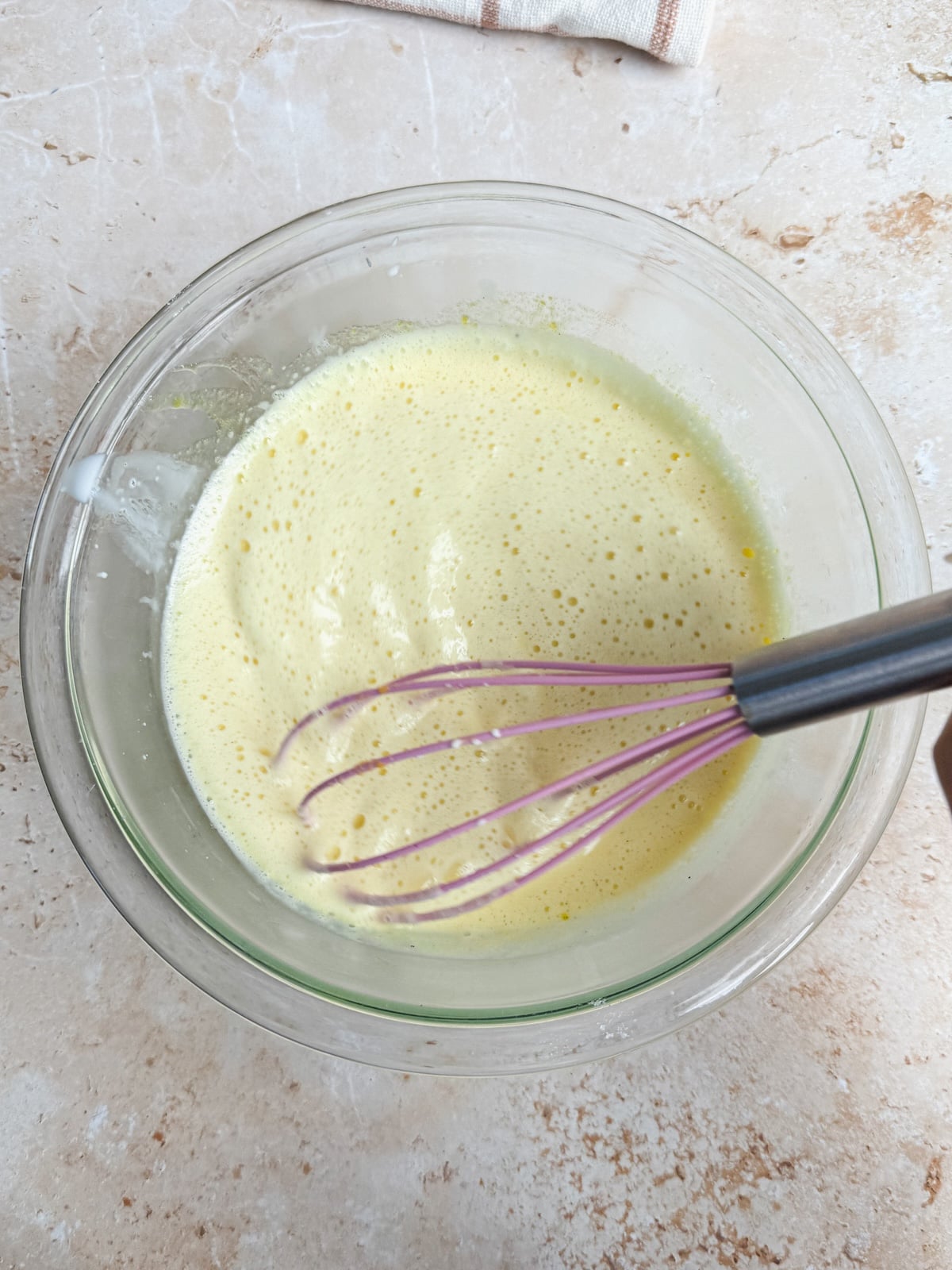 A glass bowl containing a frothy, pale yellow mixture is being whisked with a metal whisk on a light-colored countertop.