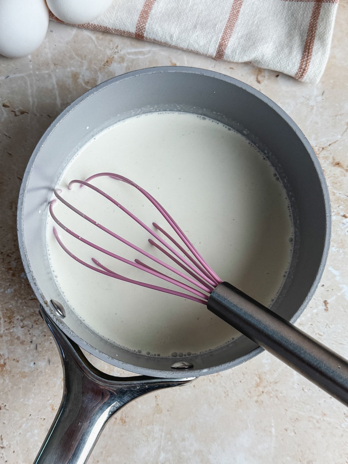 A pink whisk rests in a gray saucepan filled with cream, placed on a light-colored countertop next to a striped kitchen towel and two eggs.