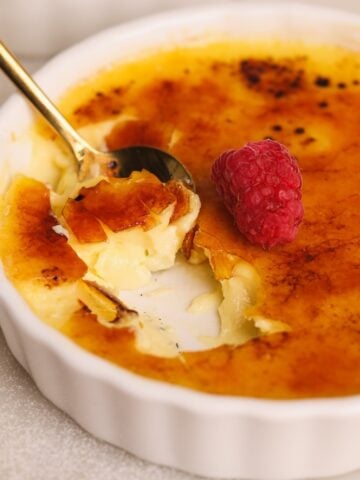 A close-up of crème brûlée in a white ramekin, with a spoon breaking through the caramelized sugar crust. A fresh raspberry sits on top of the dessert.