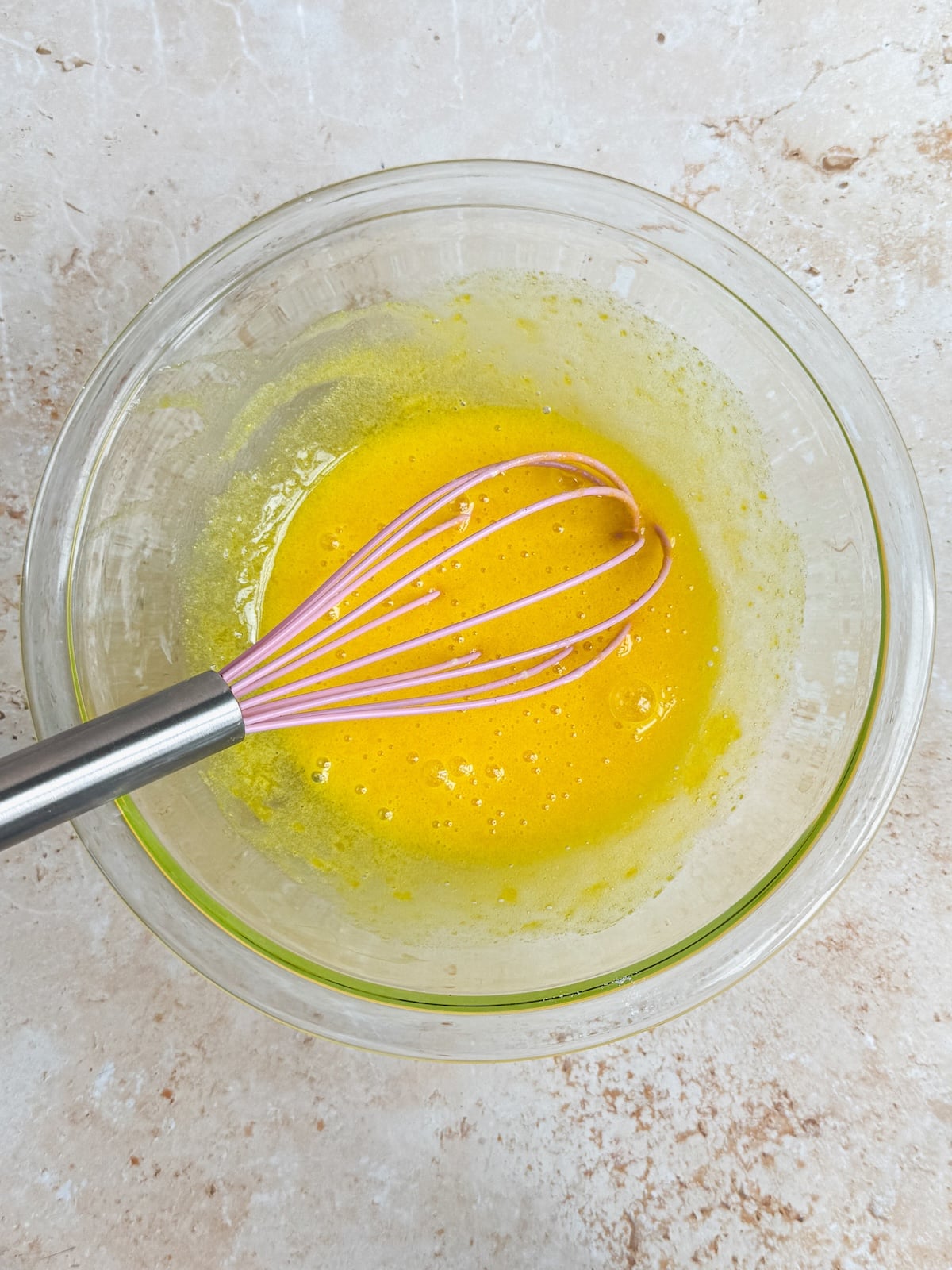 A glass bowl with a bright yellow mixture being whisked with a metal and pink whisk, placed on a light-colored countertop.