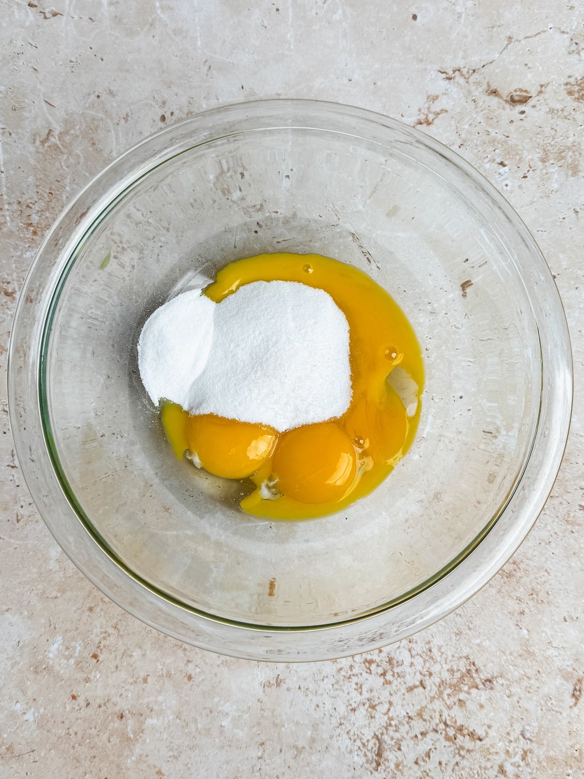 A glass bowl containing egg yolks and a mound of white sugar, sitting on a light-colored countertop.