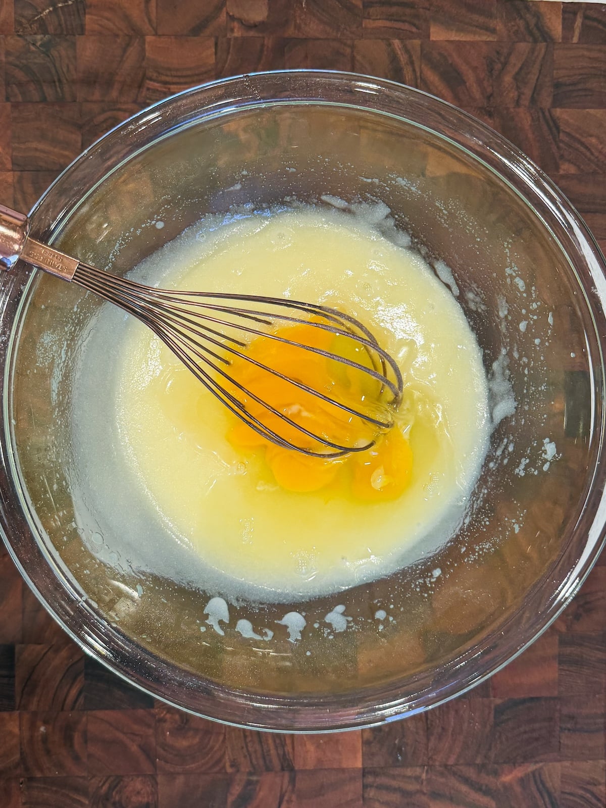 A glass bowl on a wooden surface contains melted butter and two raw eggs being mixed with a metal whisk.