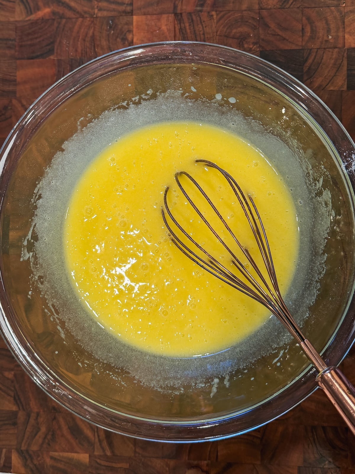 A glass bowl with a yellow liquid mixture being whisked, sitting on a wooden surface. A metal whisk is partially submerged in the mixture, indicating that it is being mixed or blended.