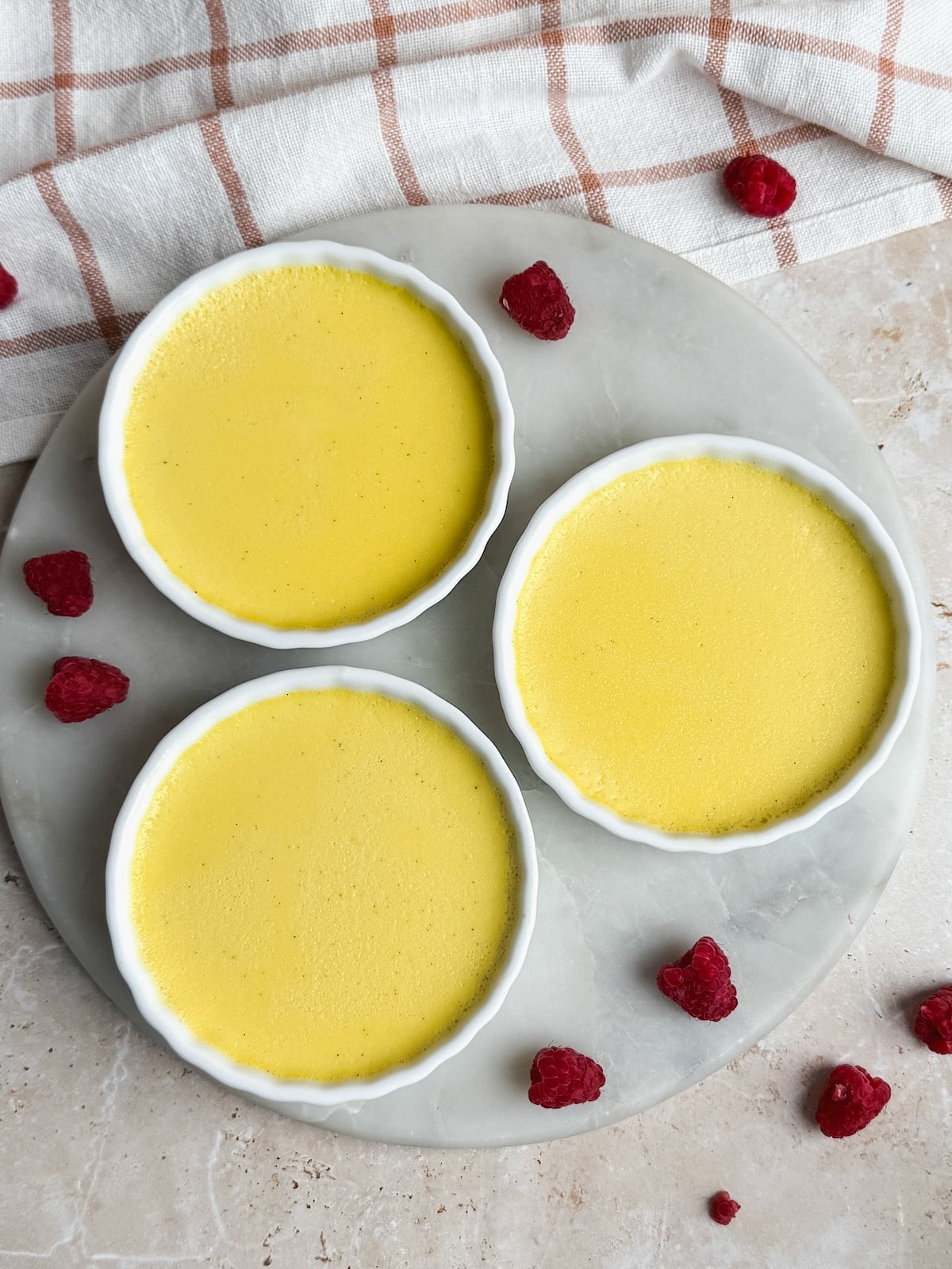 Three small round white ramekins filled with baked yellow custard are arranged on a light marble surface, surrounded by scattered raspberries. A beige and white checked cloth is partially visible in the background.