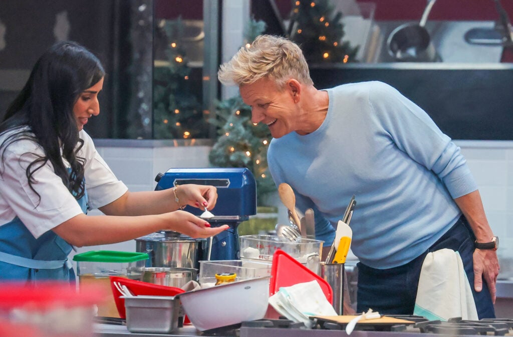 A woman in a chef’s uniform shows a dish to a smiling man in a light blue sweater in a busy kitchen, with various kitchen tools and ingredients on the counter.