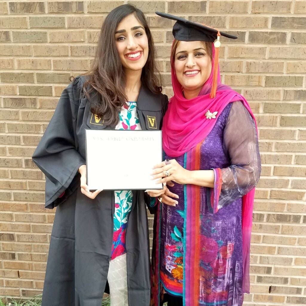 Two women smiling in front of a brick wall. One woman is wearing a graduation cap and gown, holding a diploma. The other woman is dressed in a colorful traditional outfit with a pink scarf.
