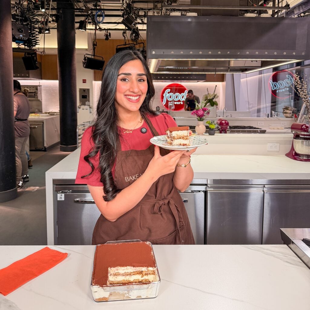 A smiling woman in a brown apron holds a plate with a slice of tiramisu in a professional kitchen studio. A larger dish of tiramisu sits on the counter, and Food Network signs are visible in the background.