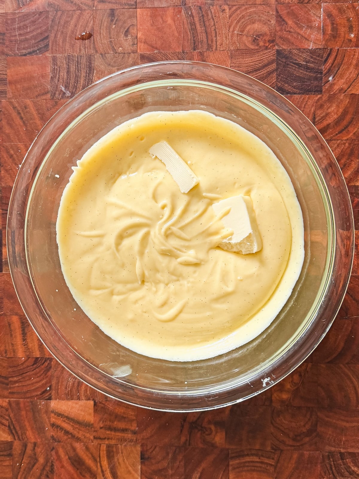 A glass bowl filled with creamy yellow custard, topped with two small pieces of butter, sits on a wooden countertop with a geometric grain pattern.