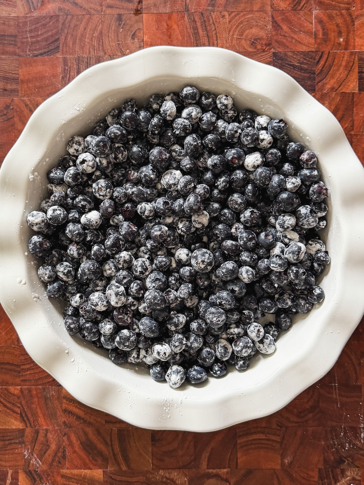 A white pie dish filled with blueberries coated in cornstarch sits on a wooden surface.