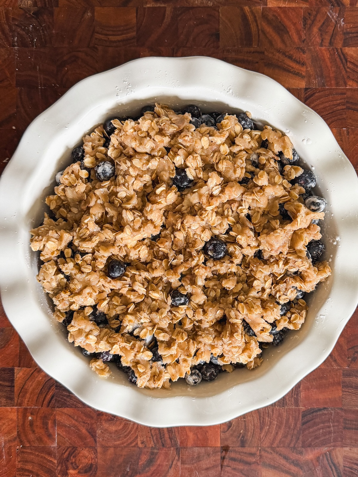 A round white pie dish filled with blueberry crisp, topped with a golden oat crumble, sits on a wooden surface. The blueberries and oat topping are visible before baking.