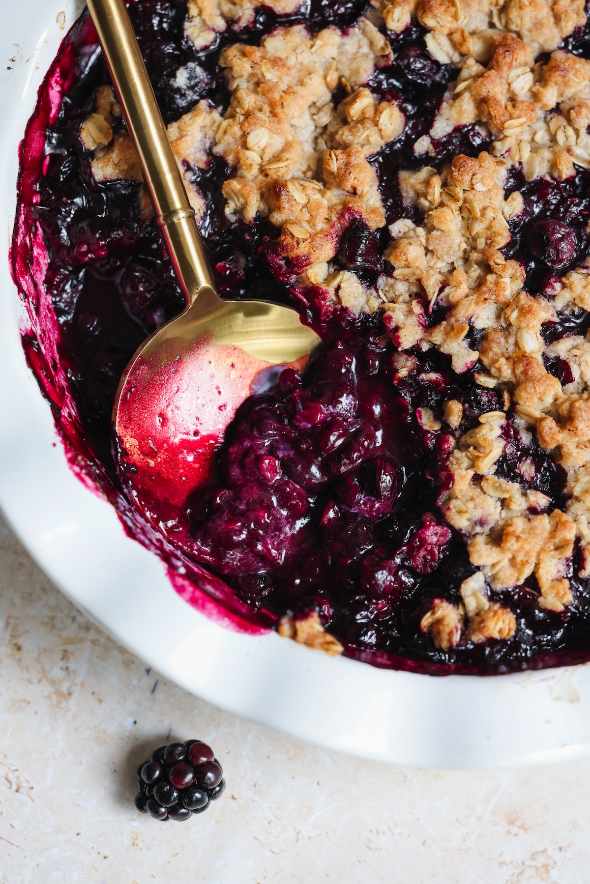 A close-up of a blueberry crumble in a white dish with a golden serving spoon, showing the dark berry filling and crumbly oat topping. A fresh blackberry sits on the countertop nearby.