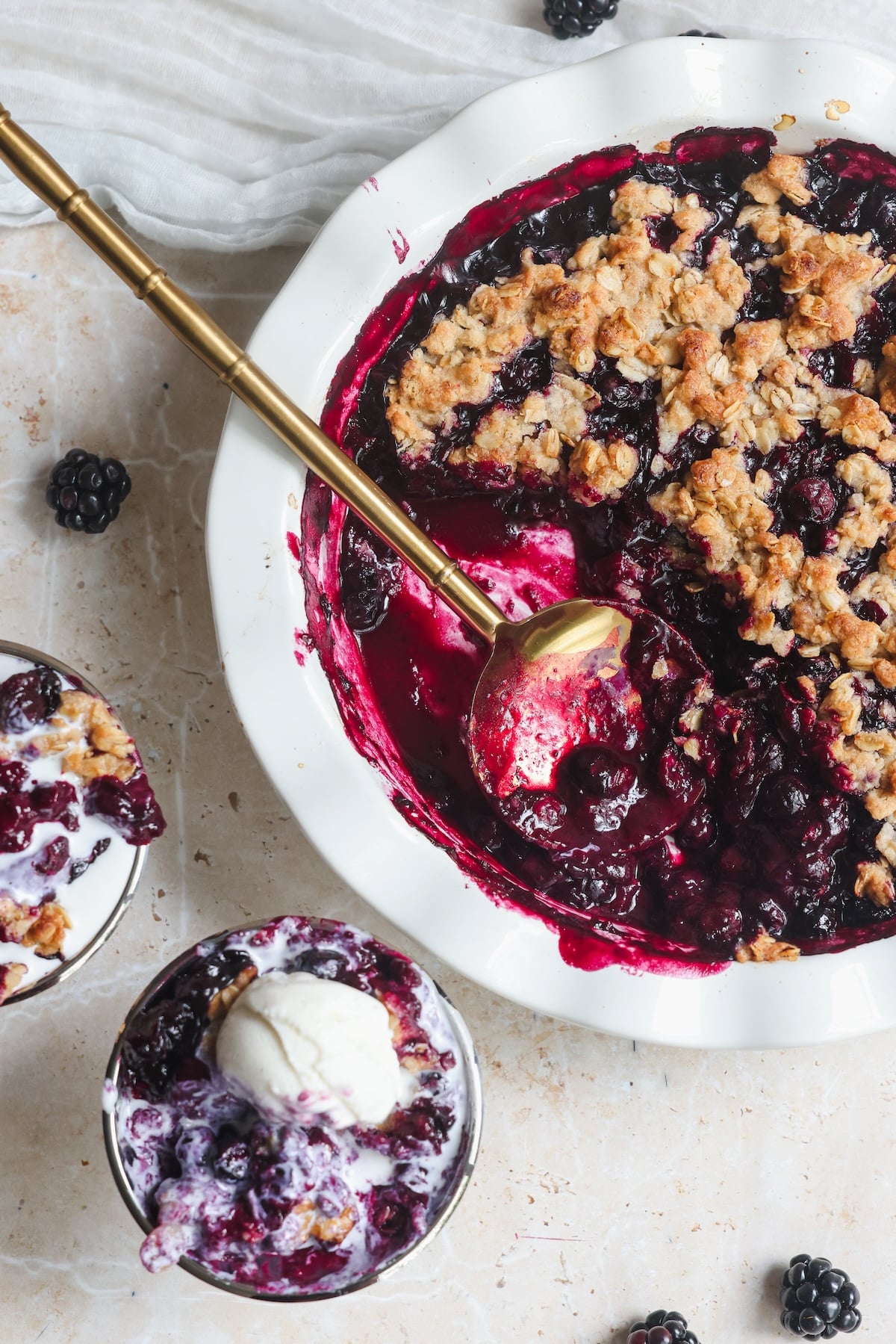 A baking dish of blueberry crisp with a golden oat topping sits beside two bowls filled with the dessert, one topped with a scoop of vanilla ice cream. A gold spoon rests in the dish; fresh berries are scattered nearby.