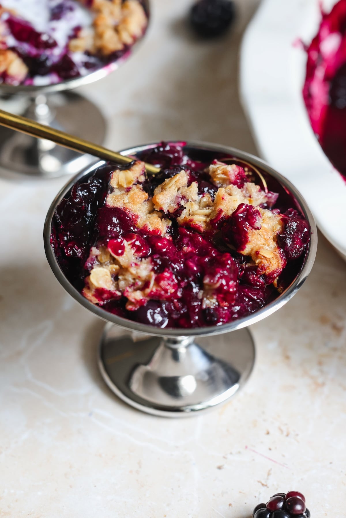 A metal dessert dish filled with a blueberry crumble topped with golden oat topping, beside scattered blackberries and a baking dish on a light countertop.