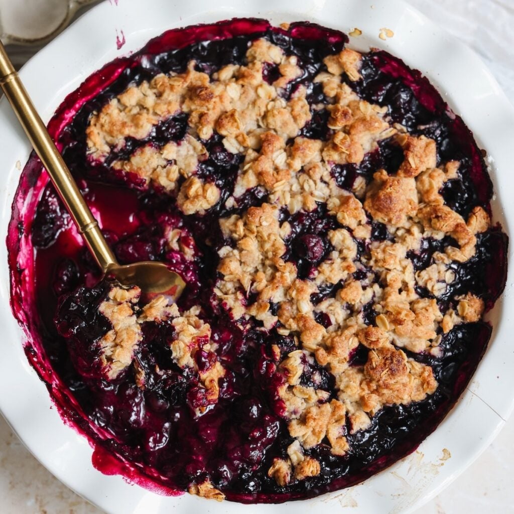 A baked blueberry crisp in a white dish with a golden oat topping and a portion scooped out, revealing the juicy, purple blackberry filling. A gold serving spoon rests in the dish.