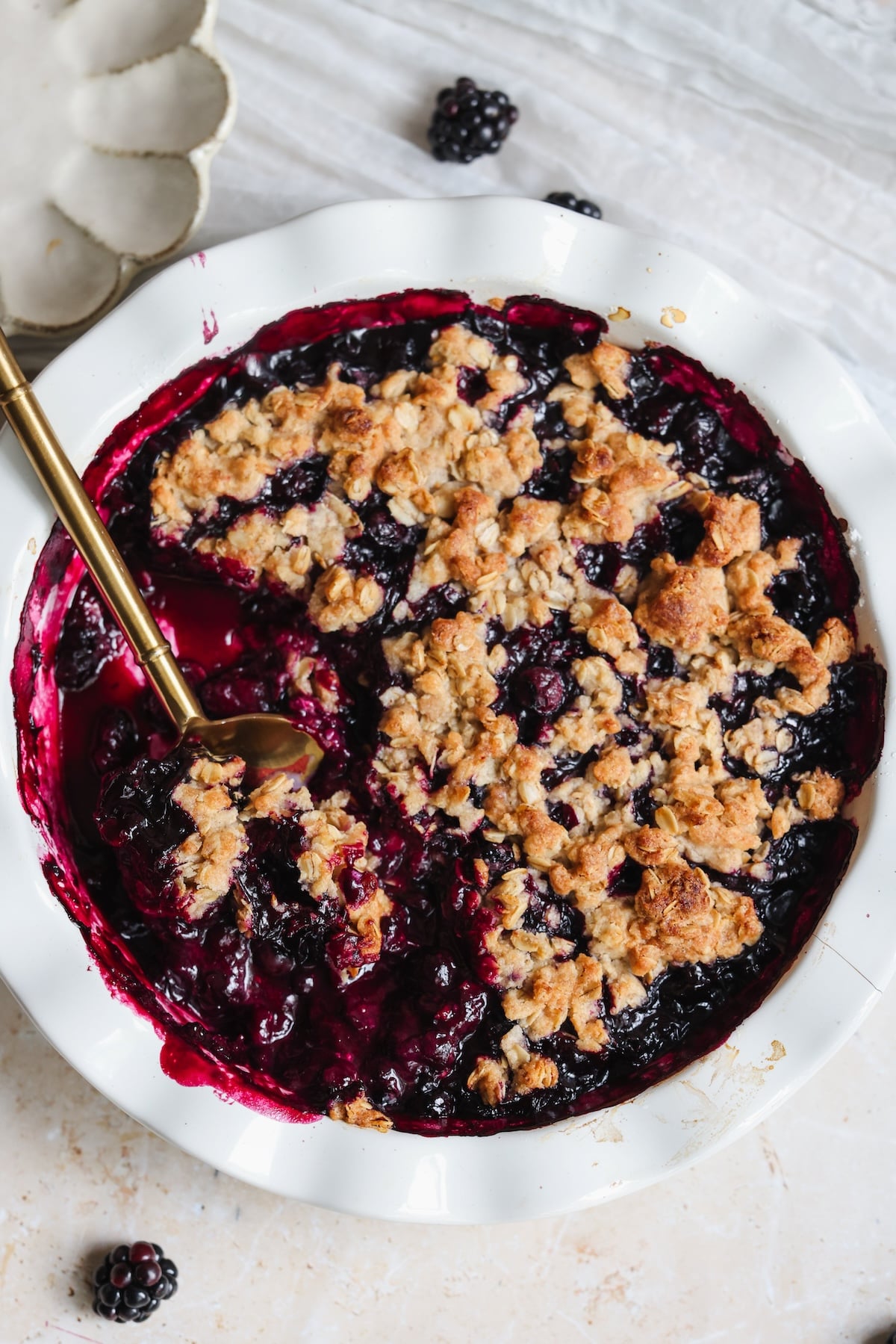 A close-up of a blueberry crumble in a white pie dish, with a golden-brown oat topping and a serving spoon in the dish.