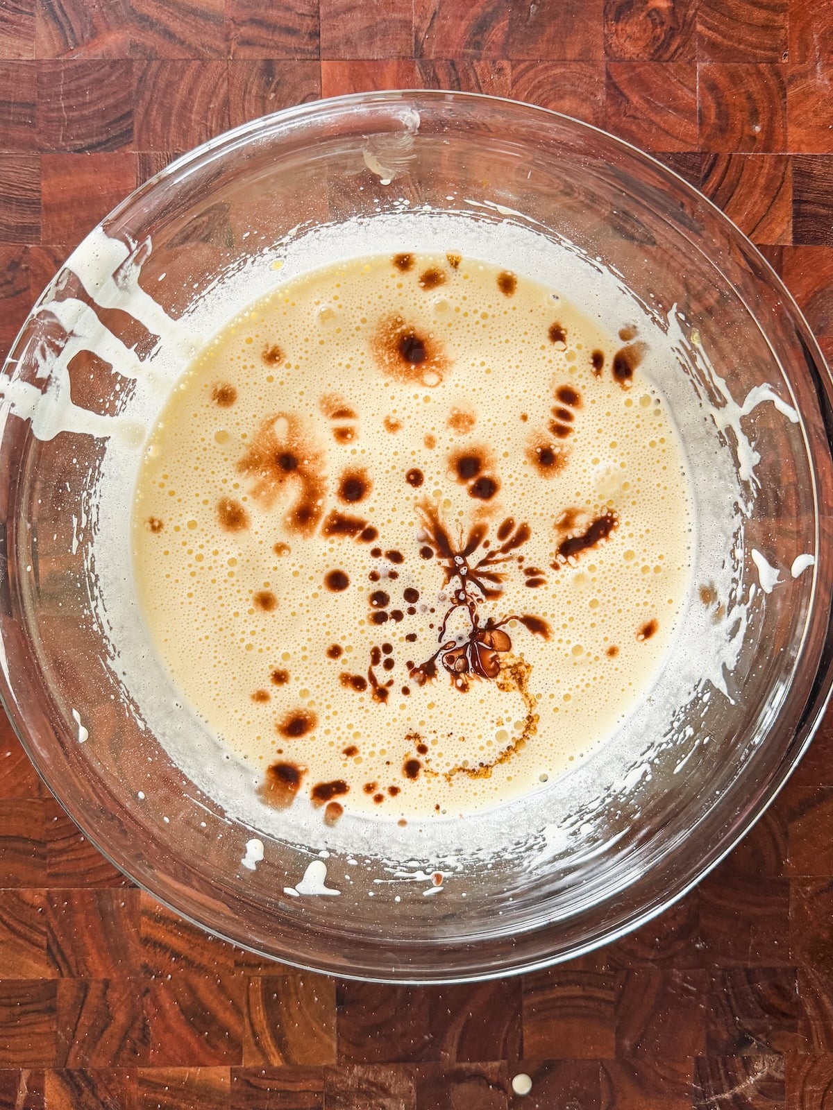 A glass bowl filled with a pale yellow batter, topped with droplets of vanilla extract, sits on a wooden surface. Some batter is splattered on the sides of the bowl.